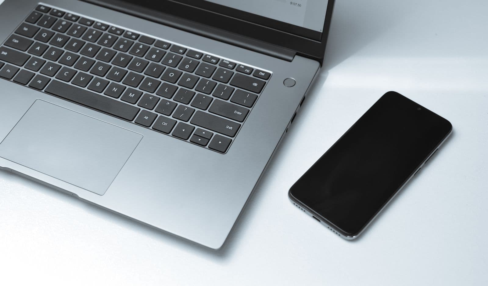 Silbernes Notebook mit Tastatur und schwarzem Smartphone auf einem weißen Tisch